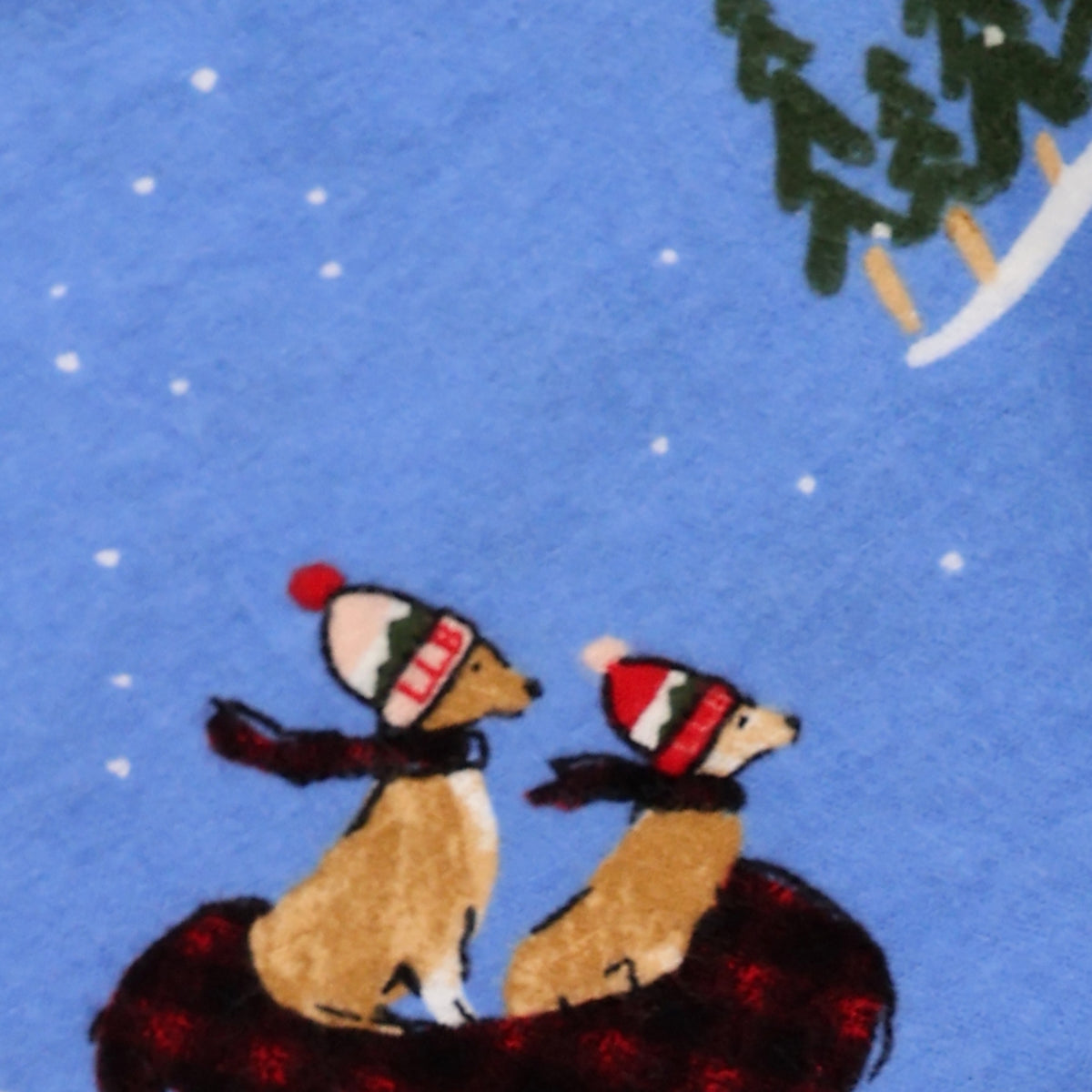 A close-up of a blue fabric with a printed winter scene featuring two dogs wearing red and green striped hats and scarves, sitting on a dark red blanket under falling white snow.