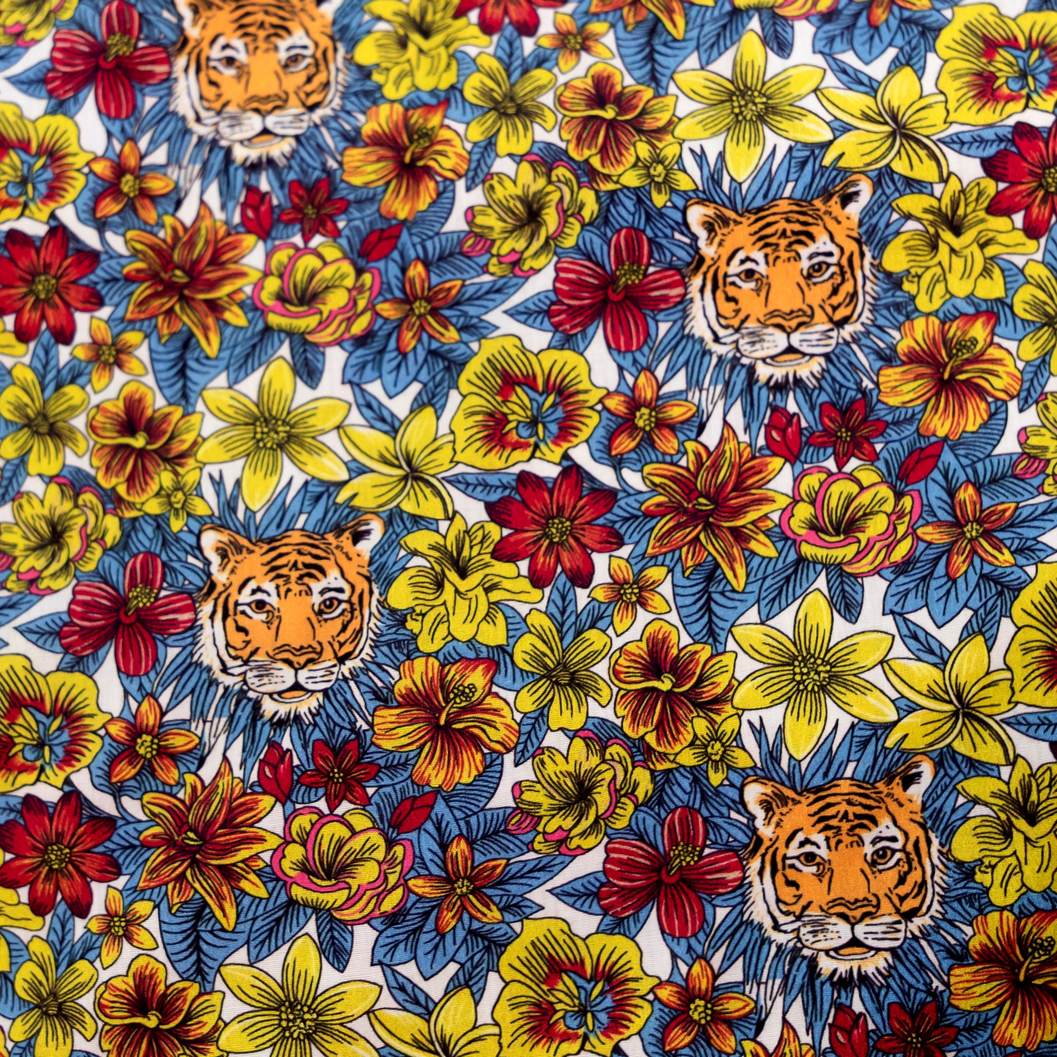 A close-up of a colorful fabric featuring a repeating pattern of three orange and black tiger faces surrounded by yellow, red, and blue tropical flowers on a white background.